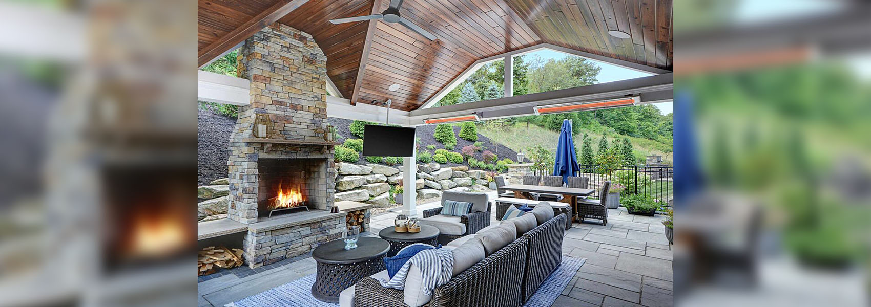 Outdoor living room with a covered gazebo, fireplace, outdoor TV, patio furniture, outdoor ceiling fan, and two electric patio heaters mounted to the frame of the gazebo