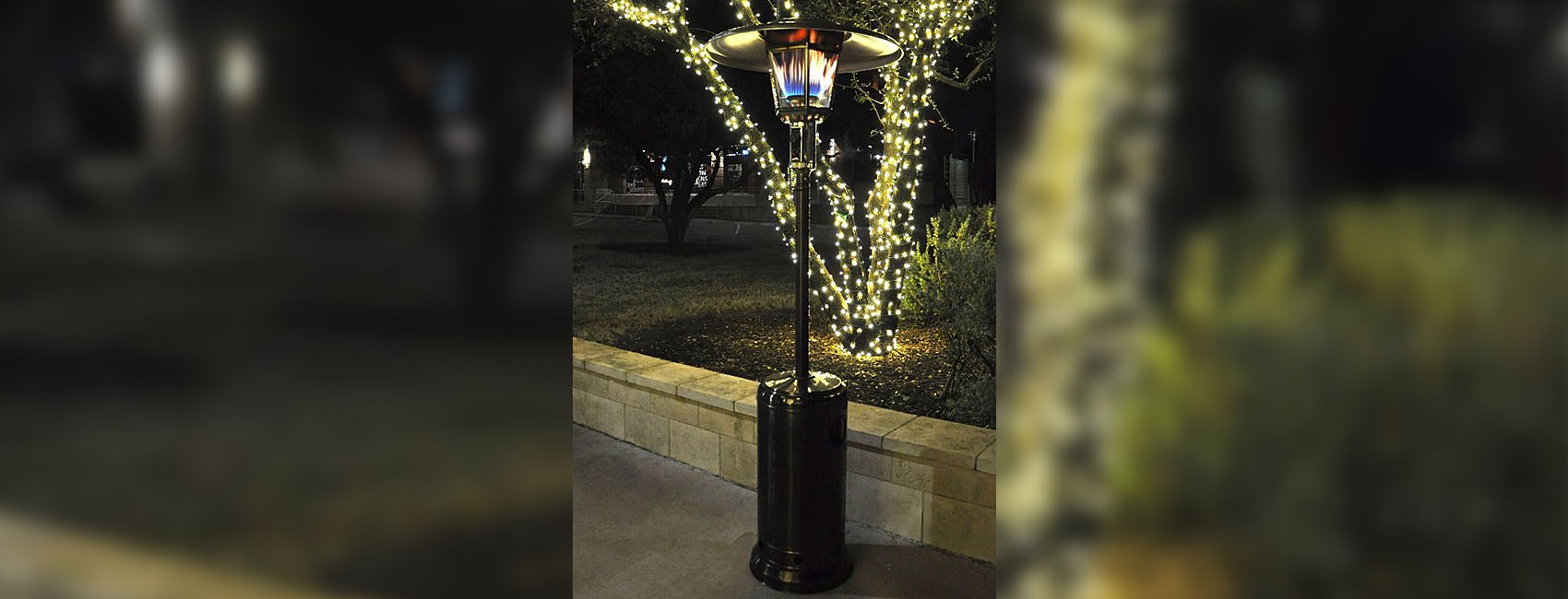 Outdoor walkway scene at night with travertine tile half-wall, greenery in the background including a tree whose trunks are lit by LED lighting, and a RADtec Real Flame RF2-MT-ANT-BRZ propane heater in the foreground
