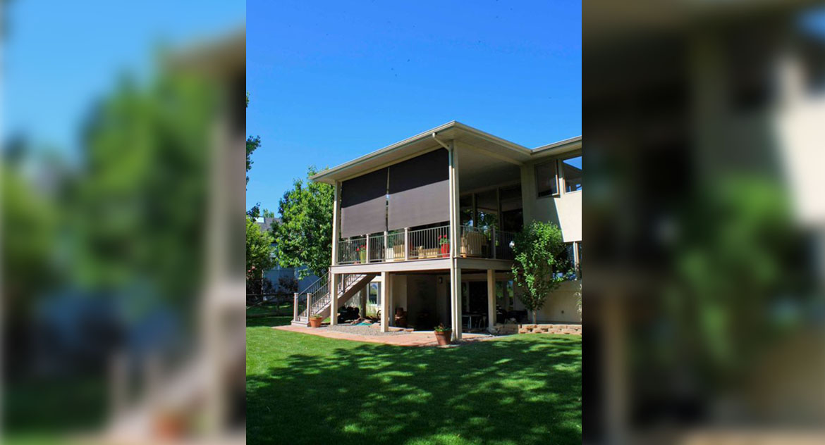 Shown from the backyard, scene of second story residential patio with metal railings, potted flowers, wicker patio furniture and two Insolroll outdoor roller shades cooling the patio