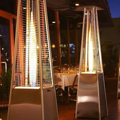 Outdoor patio scene in a restaurant, with two RADtec Tower Flame TF1-WK-BLK-GRY propane patio heaters in the foreground, with outdoor dining set with white linen tablecloths, rattan furniture in the late evening