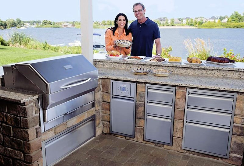a smiling couple standing behind a memphis pellet grill with a lake in the background