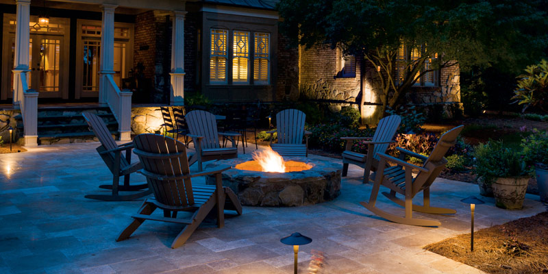 an outdoor living area with a lit fire pit and patio chairs at night with path lighting on a stone patio